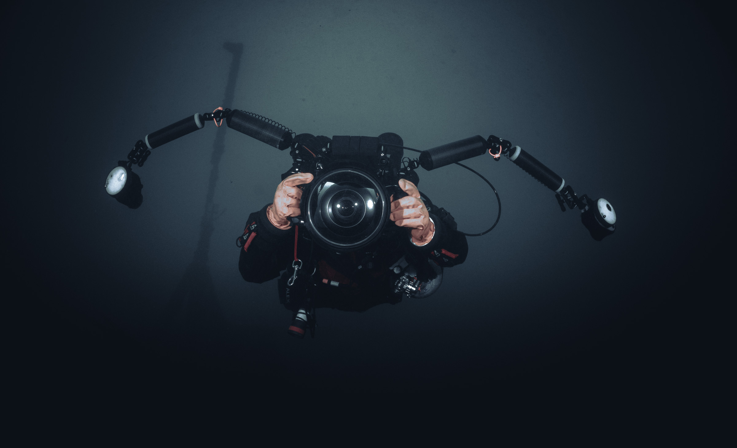 A diver holding a camera rig with a wide dome port and two strobes on arms, facing the lens