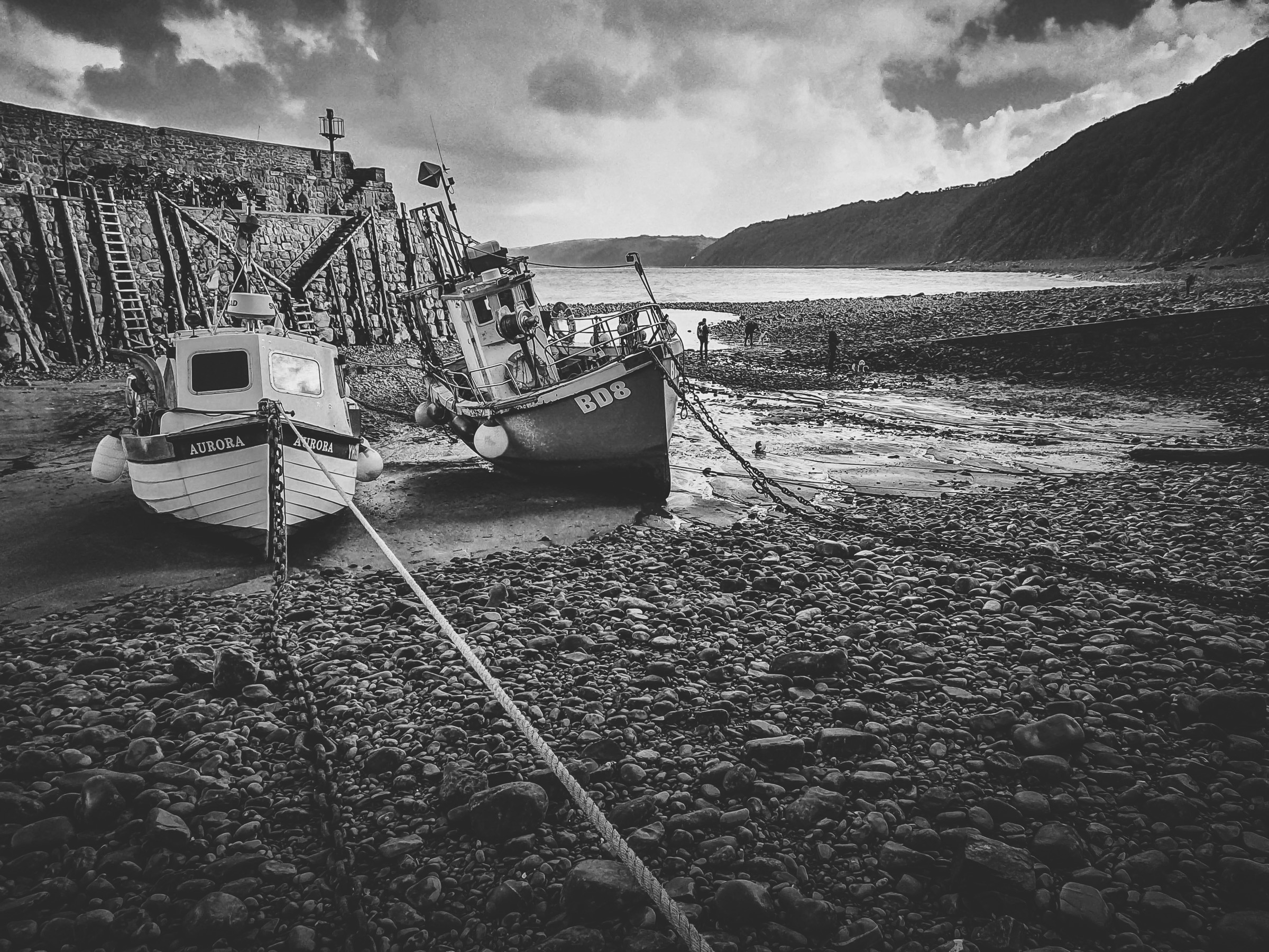 Two fishing boats beached on a pebble shore at low tide, in black and white